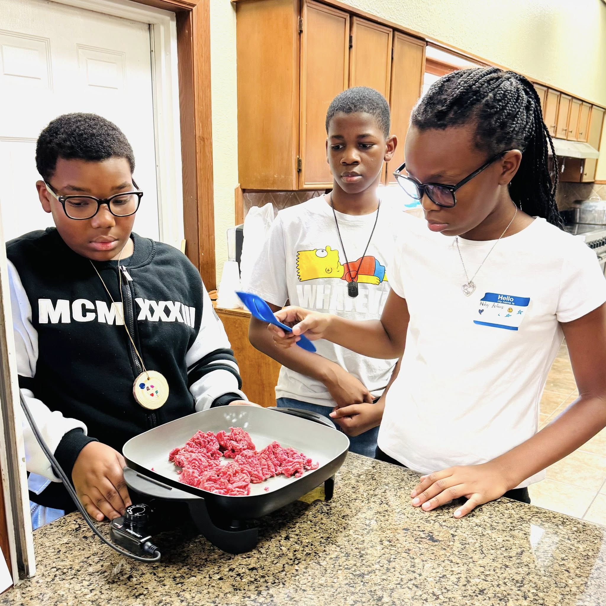 Two boys and a girl looking at raw hamburger meat in an electric skillet.